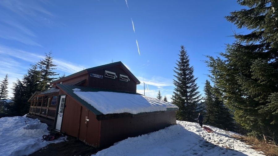 snow-covered MTTA hut surrounded by evergreen trees under a blue sky