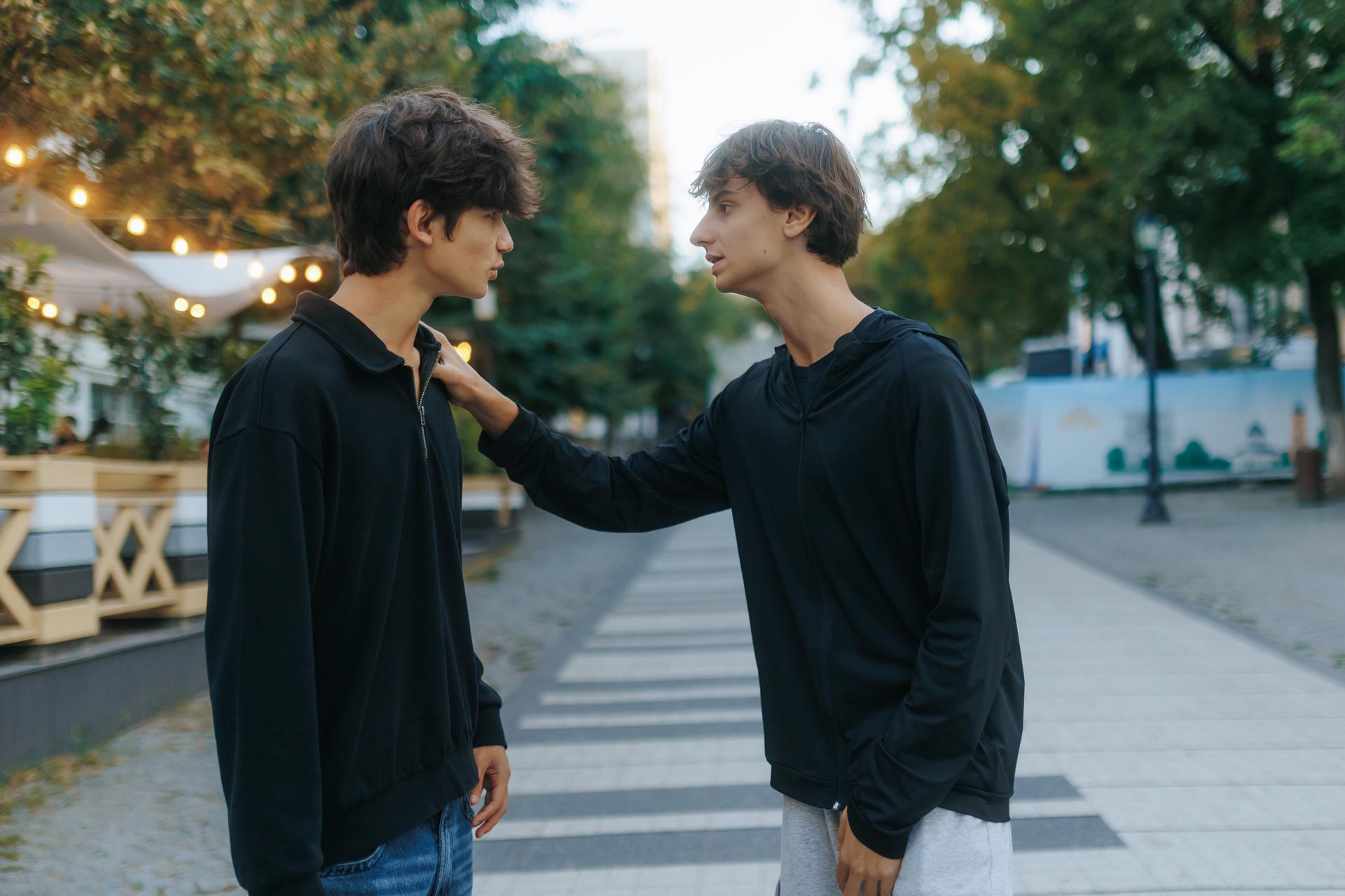 Two teens face each other on an outdoor walkway, engaged in an intense conversation. The moment shows friendship, tension, and emotions in a casual urban setting.